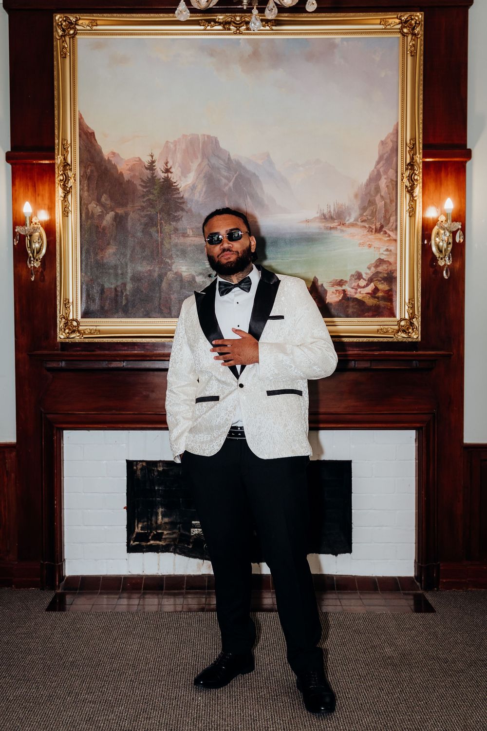 Groom in white tuxedo jacket poses before ornate painting above fireplace.