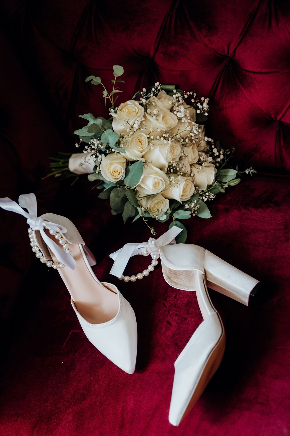 White bridal heels with pearl ankle straps beside white rose bouquet on red velvet chair.