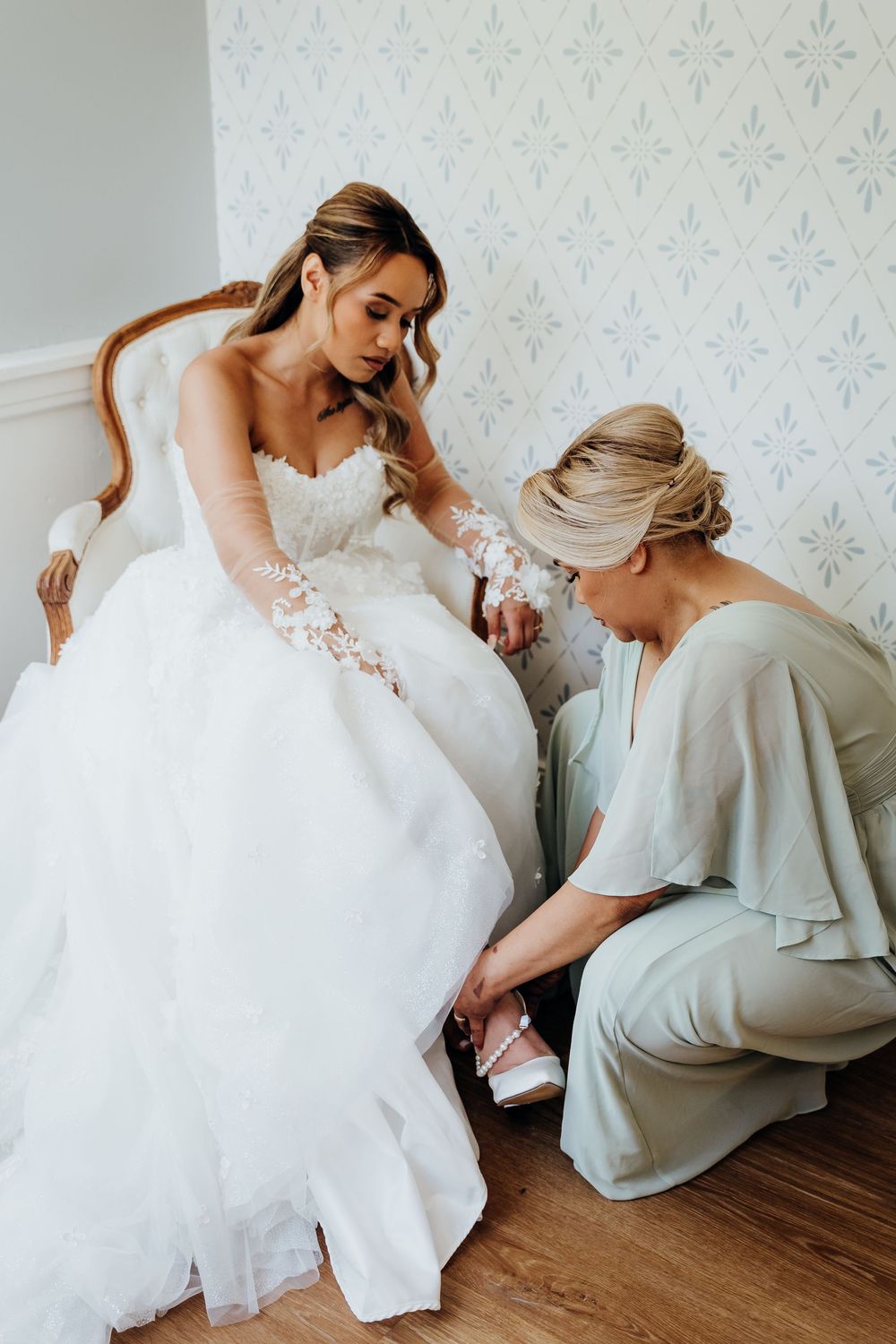 Bride in white lace gown sits on chair while bridesmaid helps put on pearl-adorned wedding heels.