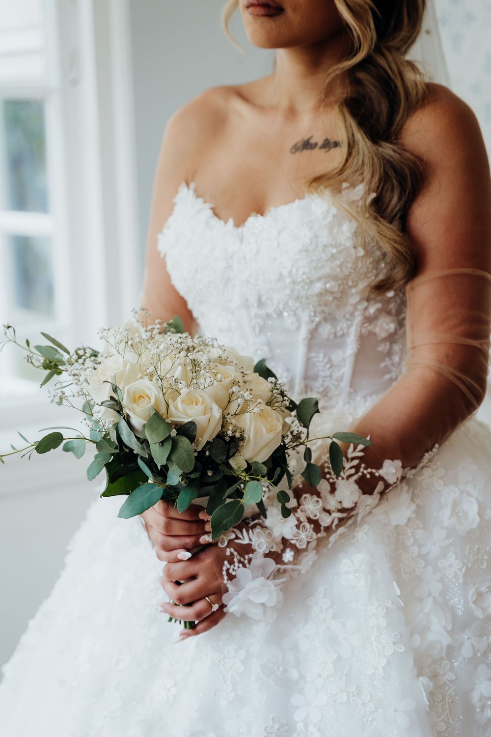Bride in strapless lace ballgown holds a white rose and eucalyptus bouquet near a bright window.