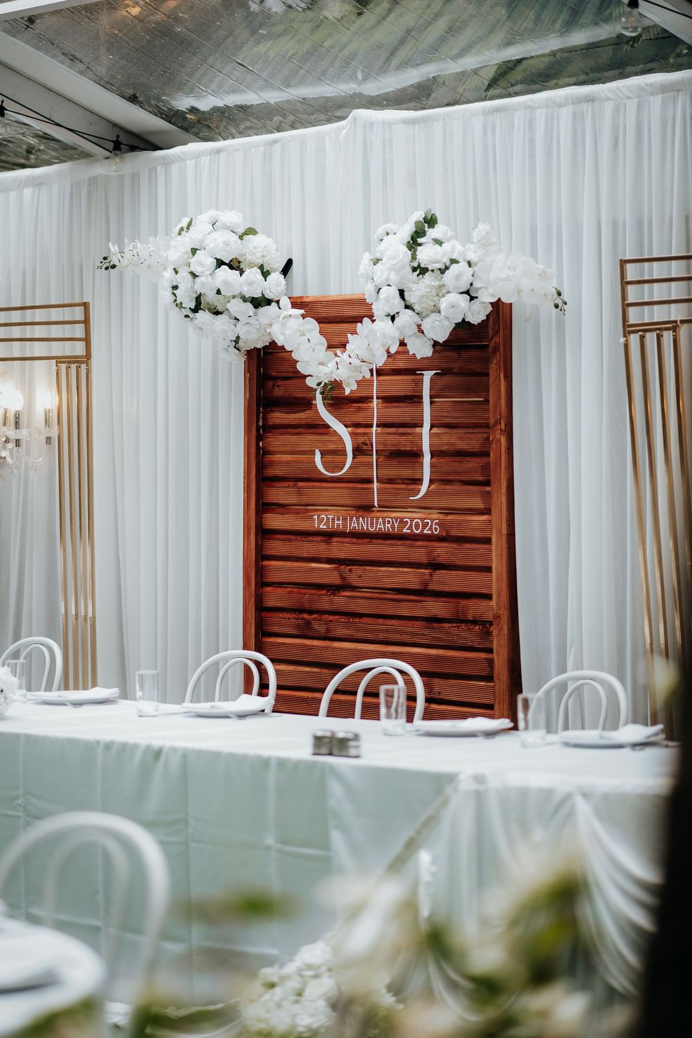 Elegant wedding head table backdrop featuring wooden panels, white floral arrangements, and monogram S&J dated 12th January 2026.
