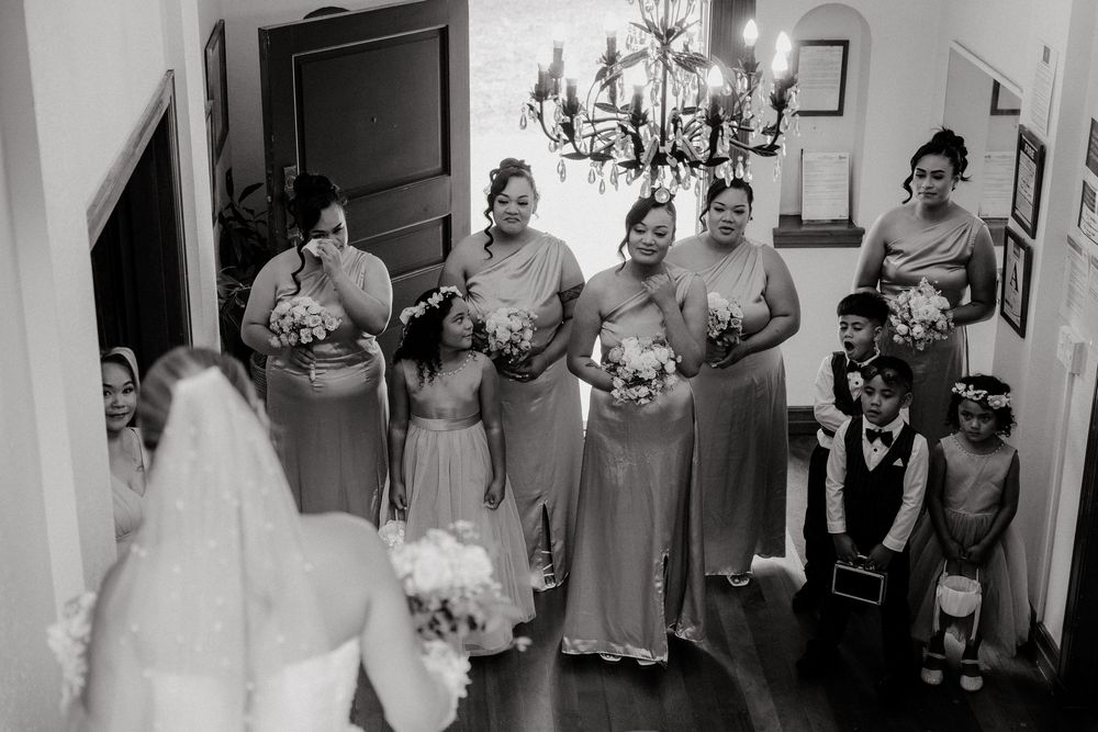 Black and white photo of bridesmaids in long gowns reacting emotionally as bride reveals her wedding dress indoors.
