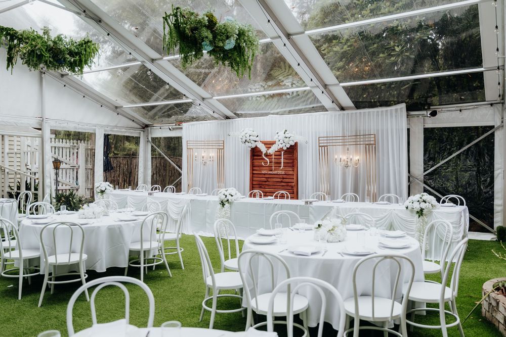 Elegant white wedding reception under clear tent with round tables, white chairs, and lush green floral arrangements.