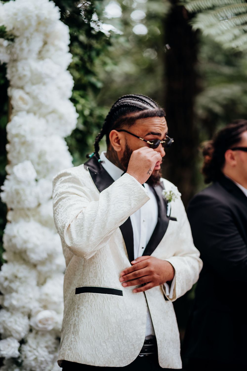 Emotional groom in white tuxedo wipes tear at outdoor wedding ceremony.