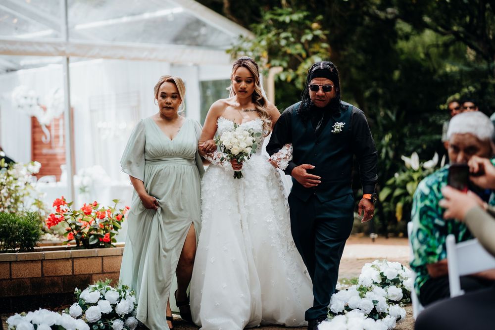 Bride in white gown walks down the aisle with bridesmaids at an outdoor wedding ceremony surrounded by white flowers.