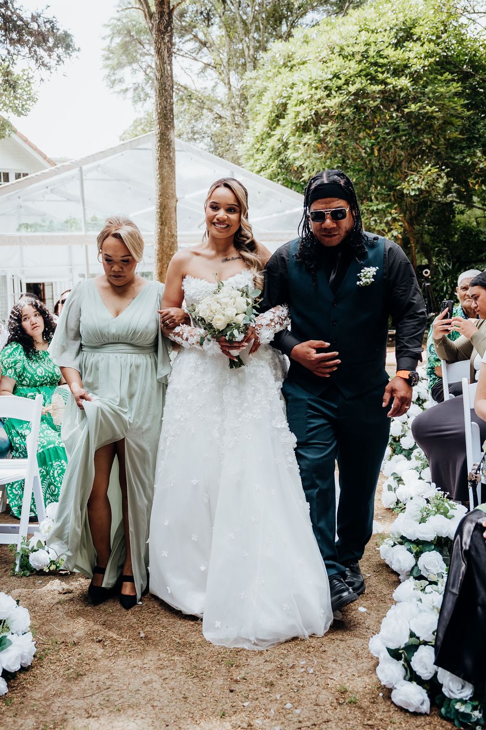 Bride in white gown walks down outdoor aisle escorted by two family members at garden wedding.