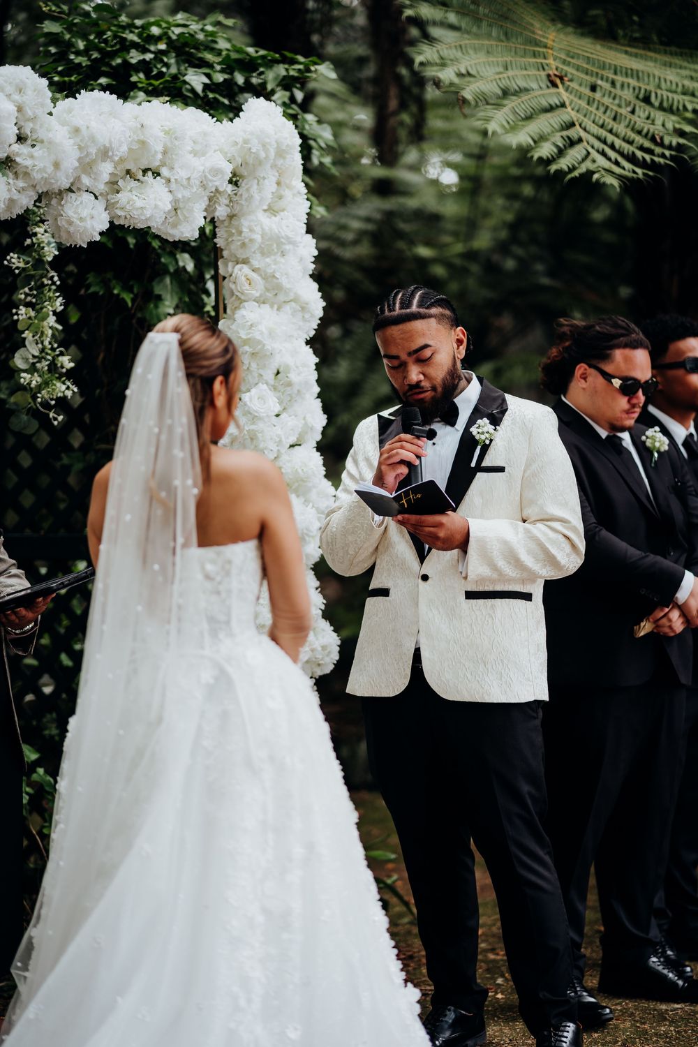 Groom in white tuxedo reads vows to bride at outdoor wedding ceremony beneath white floral arch.