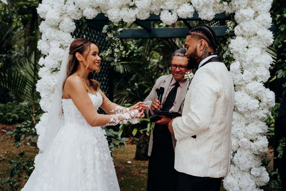A bride and groom exchange rings under a white floral arch during an outdoor wedding ceremony.