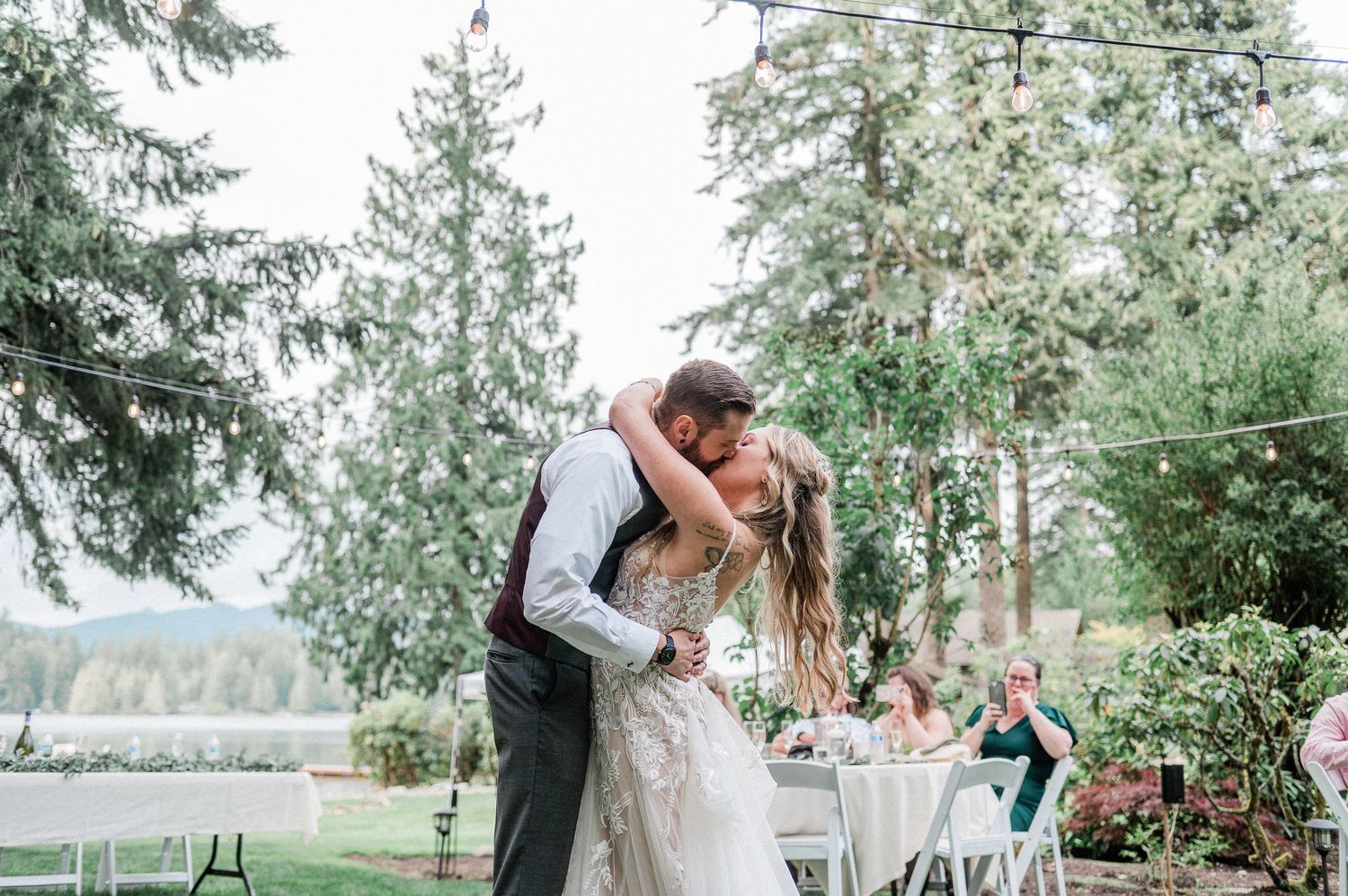 Bride and groom share a romantic kiss outdoors at backyard wedding reception surrounded by tall evergreen trees.
