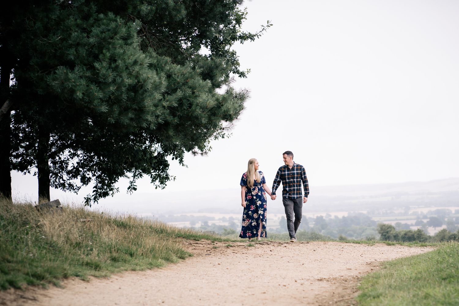 Couple walking hand in hand along a scenic dirt path beside a large tree with a countryside view.