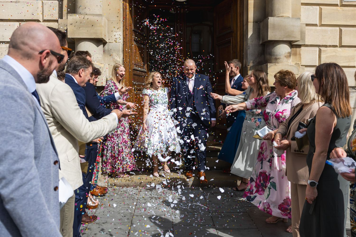 Newlyweds exit church amid confetti shower, surrounded by cheering wedding guests in elegant attire on sunny day.