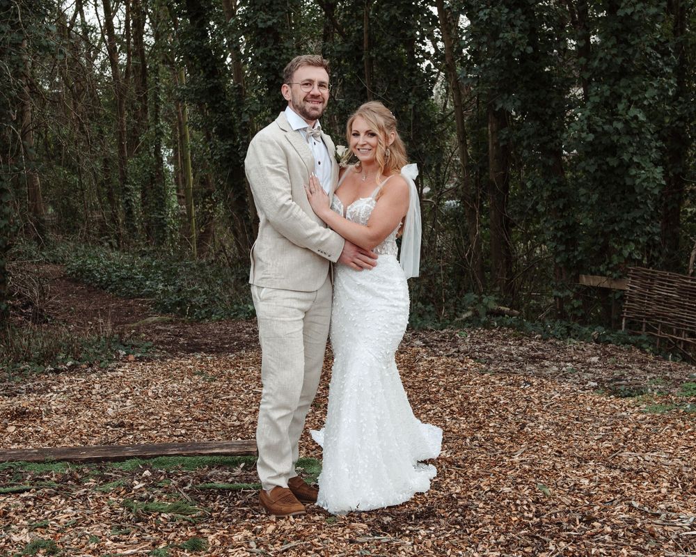 Bride and groom posing together in a woodland setting, wearing white lace gown and beige suit.
