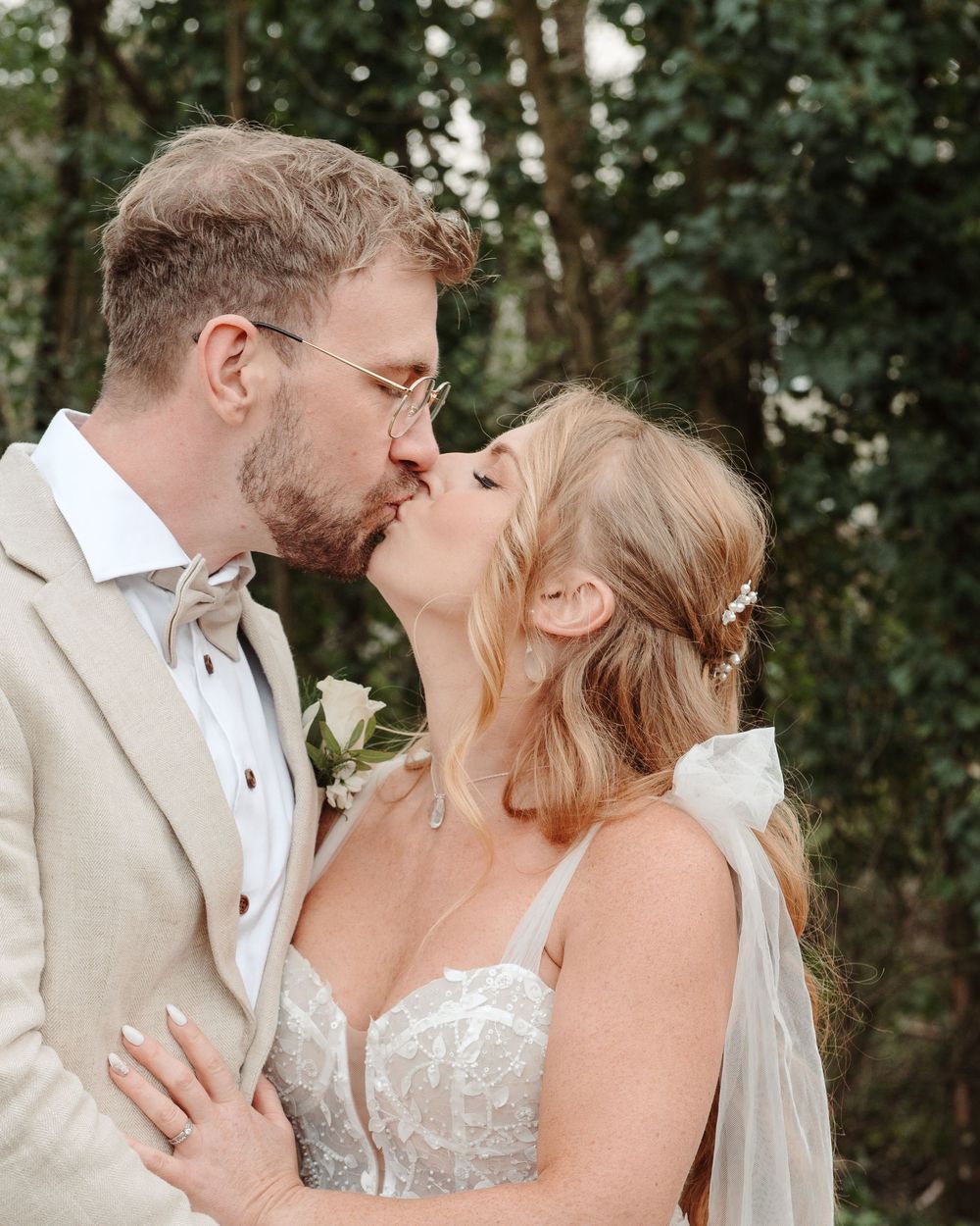 A bride and groom share a romantic kiss outdoors, she in a lace gown with veil, he in a beige suit with boutonniere.