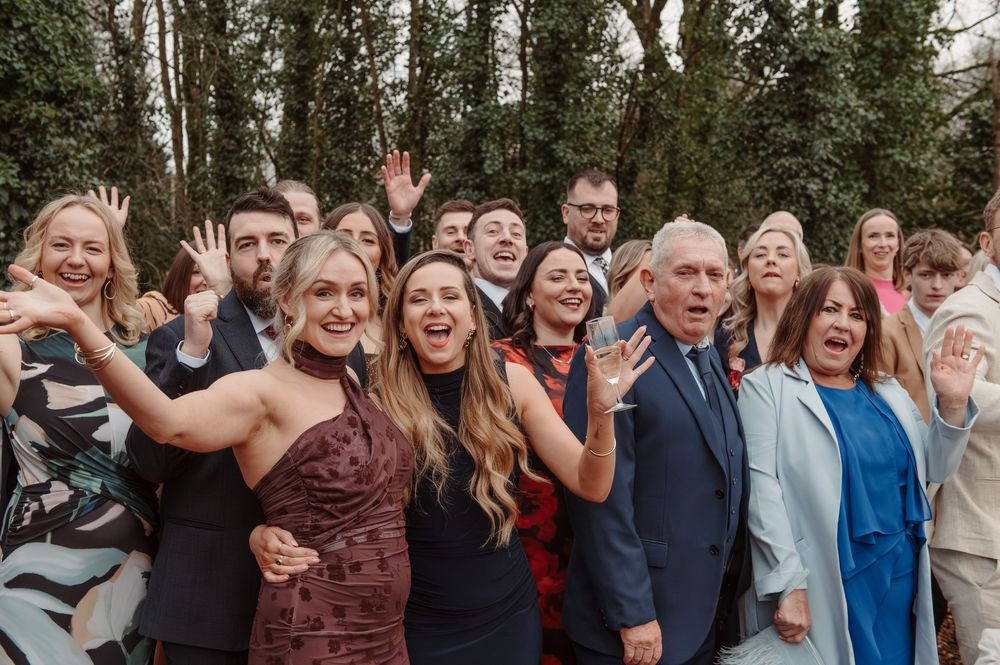 Large group of happy wedding guests posing and waving together outdoors at a celebration.