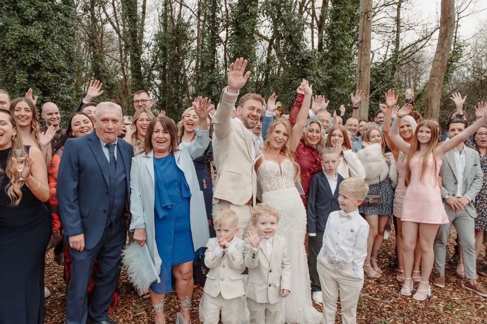 Large wedding group photo with bride and groom surrounded by smiling guests waving in a woodland setting.