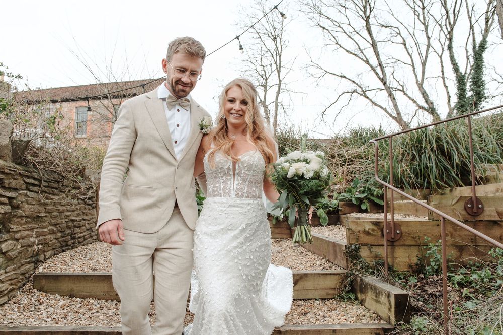 Smiling bride and groom posing together outdoors holding a bouquet in elegant wedding attire.