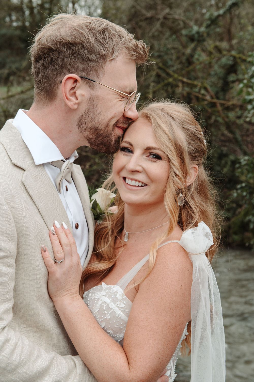 Smiling bride in lace gown embraces groom in beige suit outdoors near greenery on their wedding day.