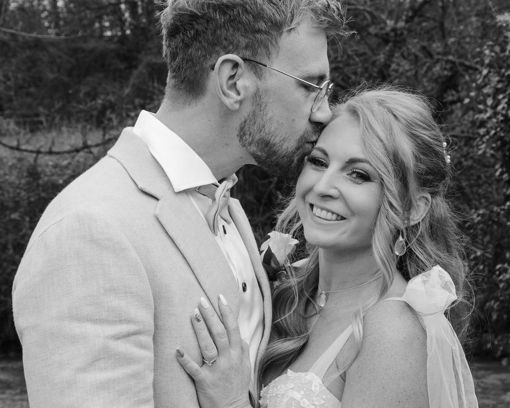 Black and white photo of a groom kissing bride on cheek, bride smiling at camera in wedding attire.