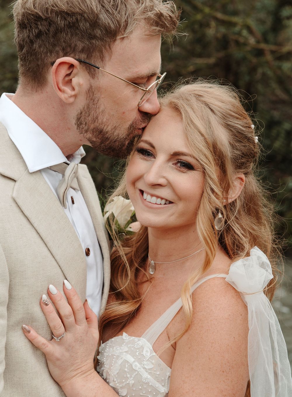 Groom kissing smiling bride on cheek, bride wearing white lace dress with floral shoulder detail.