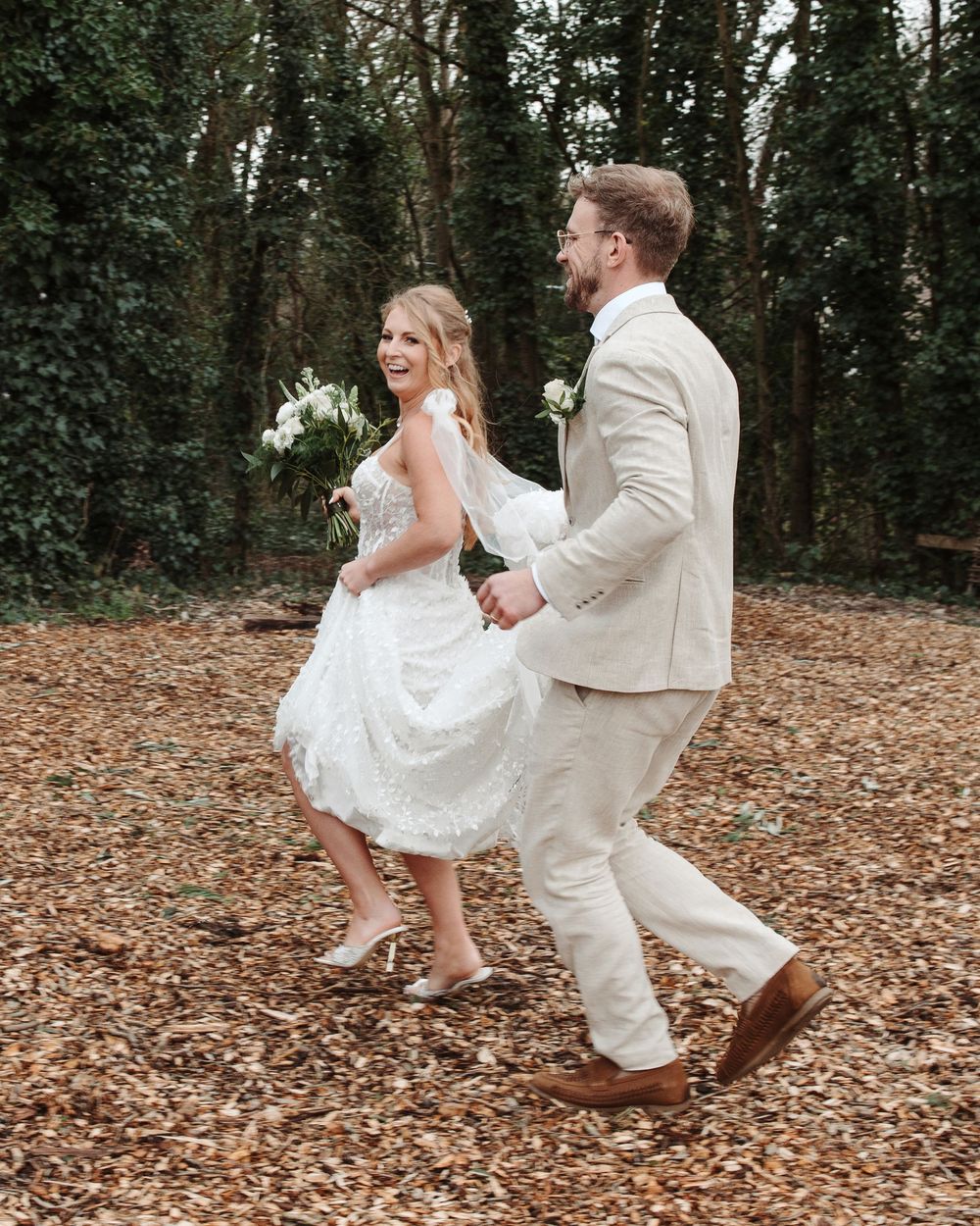 Bride and groom walking hand in hand through autumn leaves in a woodland setting on their wedding day.