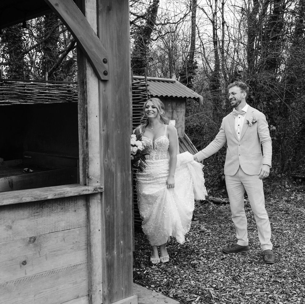 Black and white photo of bride and groom walking hand in hand near rustic wooden structure outdoors.