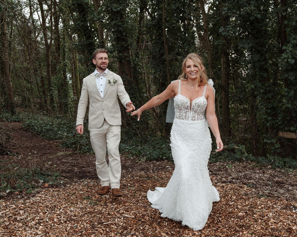 Bride and groom holding hands walking through woodland in a lace wedding dress and beige suit.