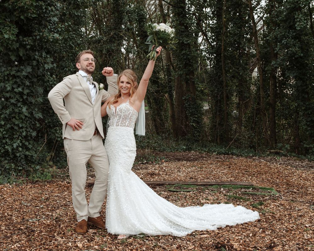 Joyful bride raising bouquet in the air with groom in woodland setting covered in autumn leaves.