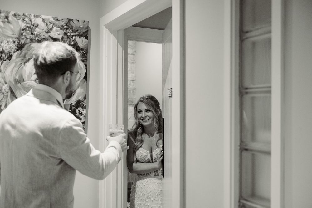 Groom opens door for smiling bride in elegant lace wedding dress during romantic first look moment.