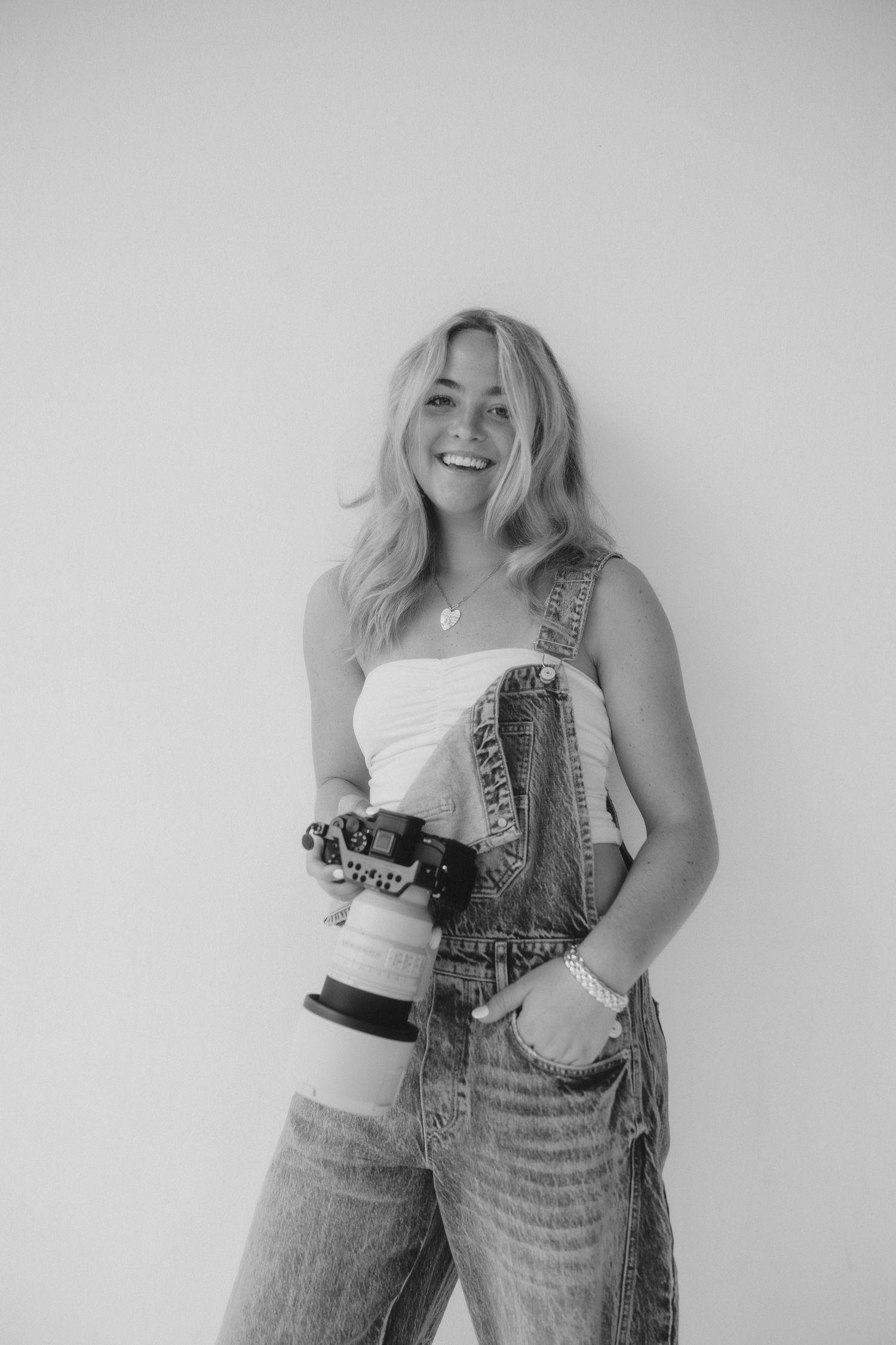 Smiling blonde woman holding a professional camera with telephoto lens, wearing denim overalls and white top, black and white photo.
