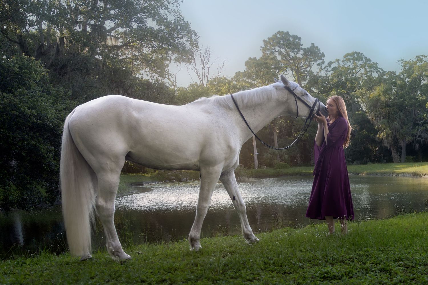 Woman in purple dress standing beside a majestic white horse near a serene pond in a forest.