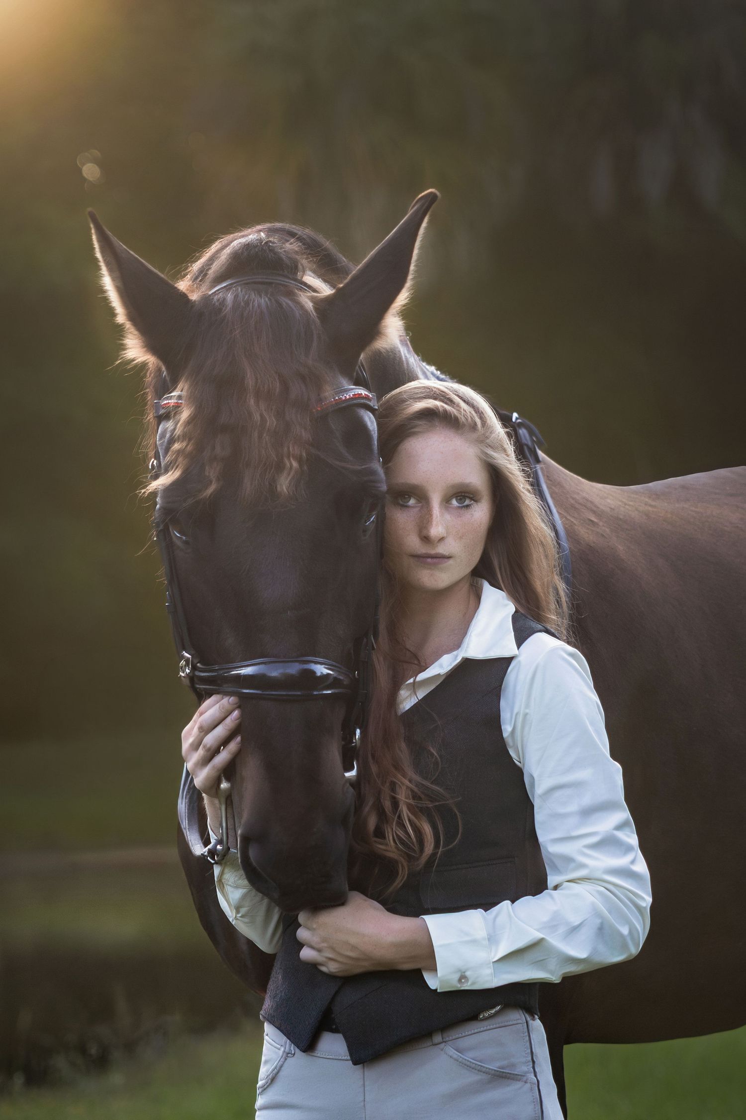 Young woman in equestrian attire embracing a dark bay horse at golden hour sunset.