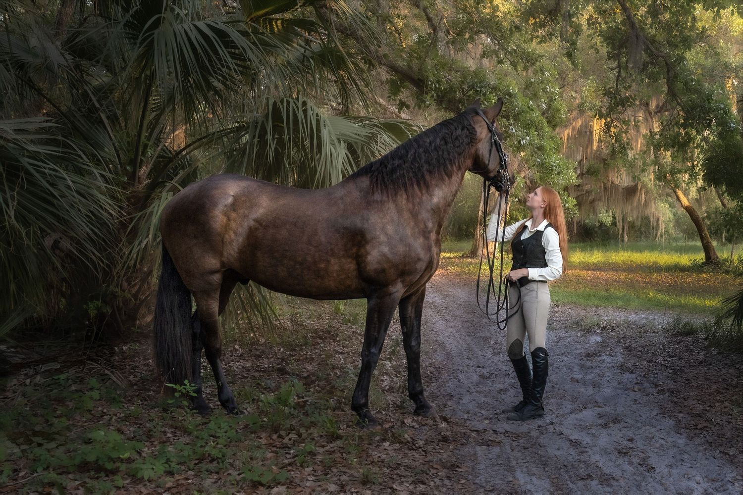 Redheaded rider in breeches standing with a dark bay horse on a wooded trail.