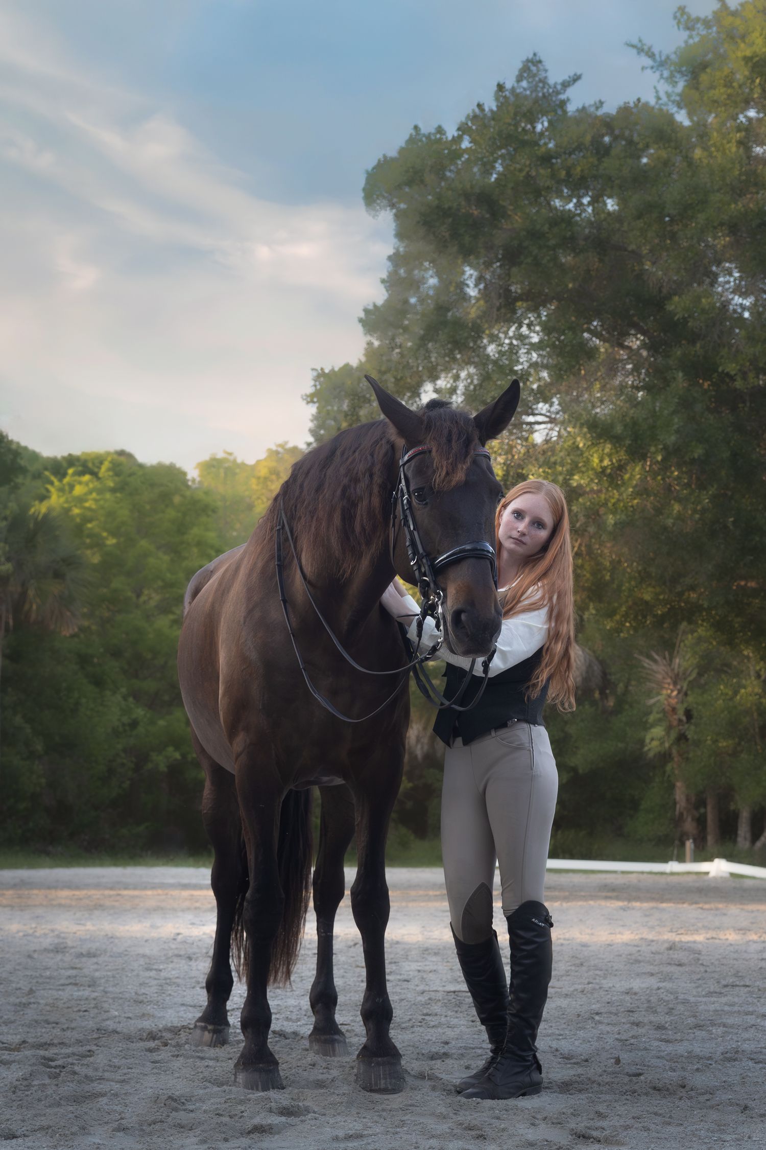 Blonde equestrian in riding boots standing beside a dark bay horse in sandy arena.
