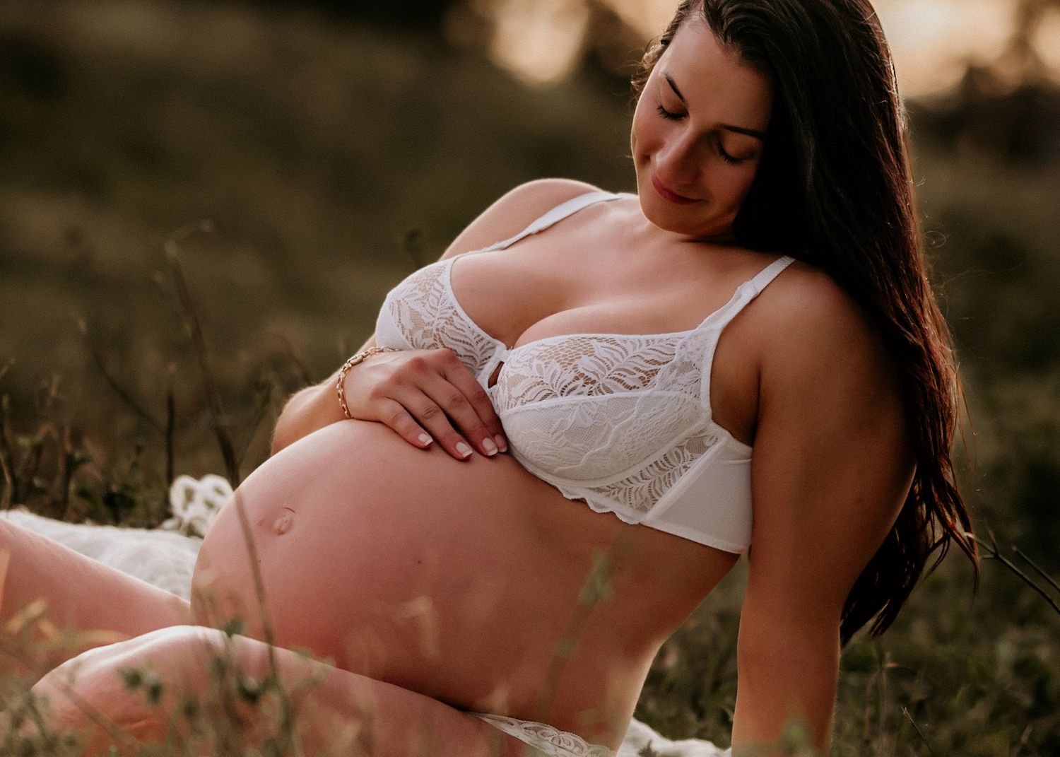 Intimate maternity portrait of expectant mother in white lingerie sitting in golden sunset field.