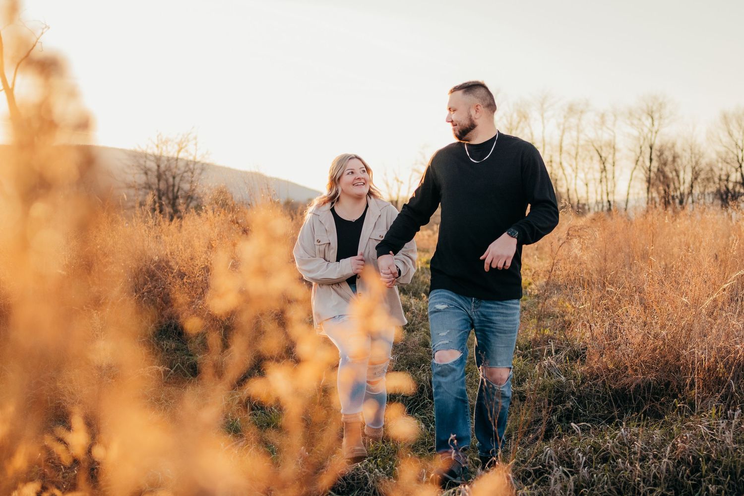 Two people walk together and laugh while surrounded by golden autumn grass and warm sunset light.