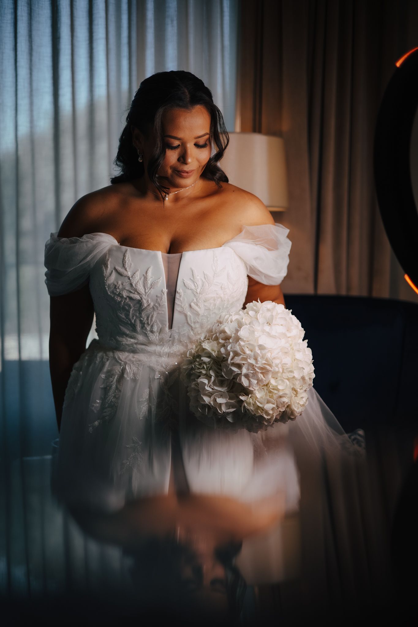A bride in an elegant off-shoulder white wedding dress with ruffled details poses in moody lighting by a window.