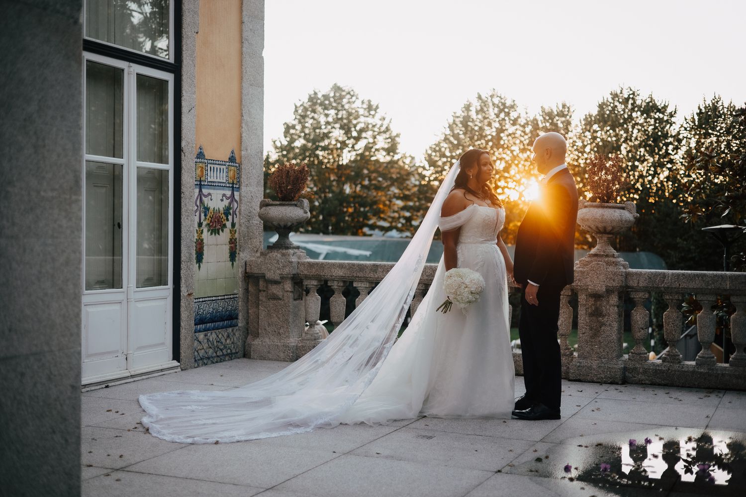 Wedding couple silhouetted against golden sunset at outdoor venue with trees and building in background.