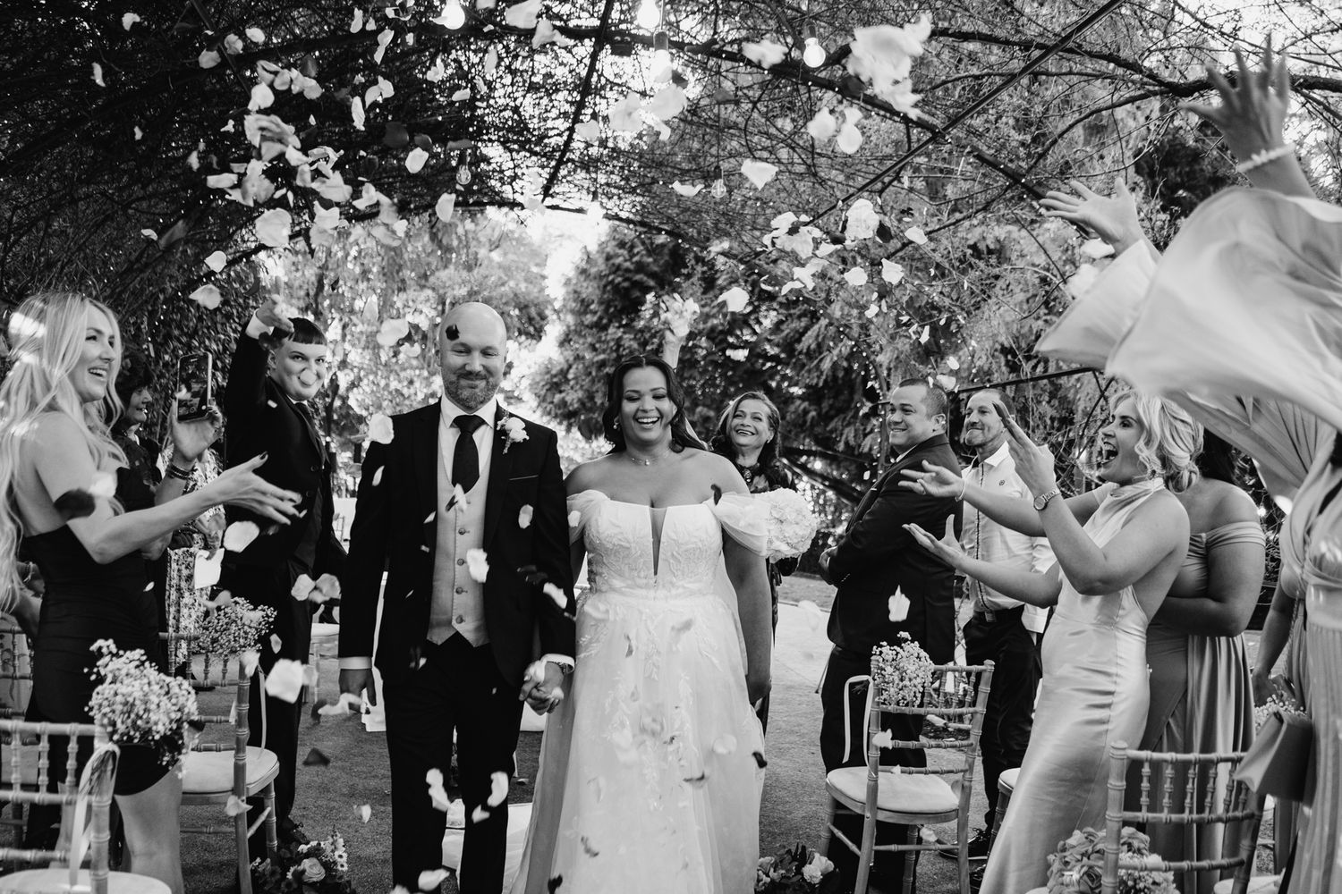 Wedding guests dance together during an outdoor reception celebration beneath string lights and trees.