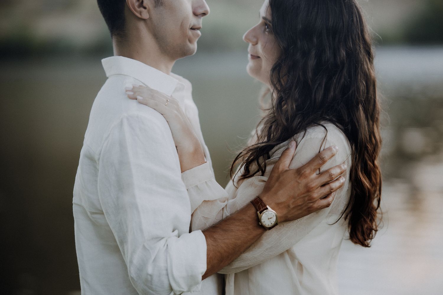 Romantic couple embracing outdoors near a lake, woman in white dress with watch, man in white shirt.