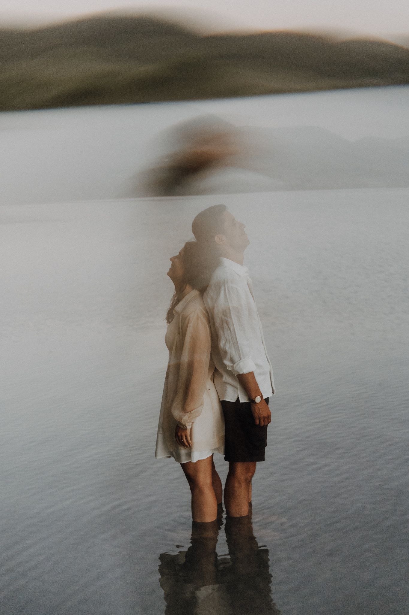 Couple standing back-to-back in shallow water, wearing white shirts, with misty hills and a serene lake in the background.