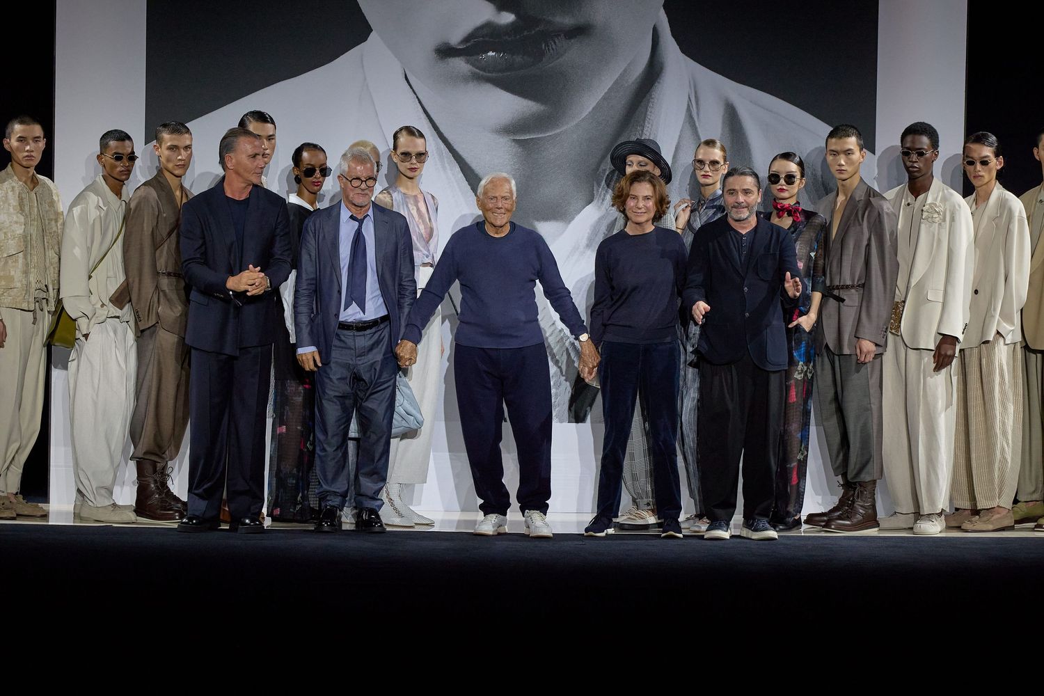 Fashion show lineup with models and designers on stage with large portrait backdrop.