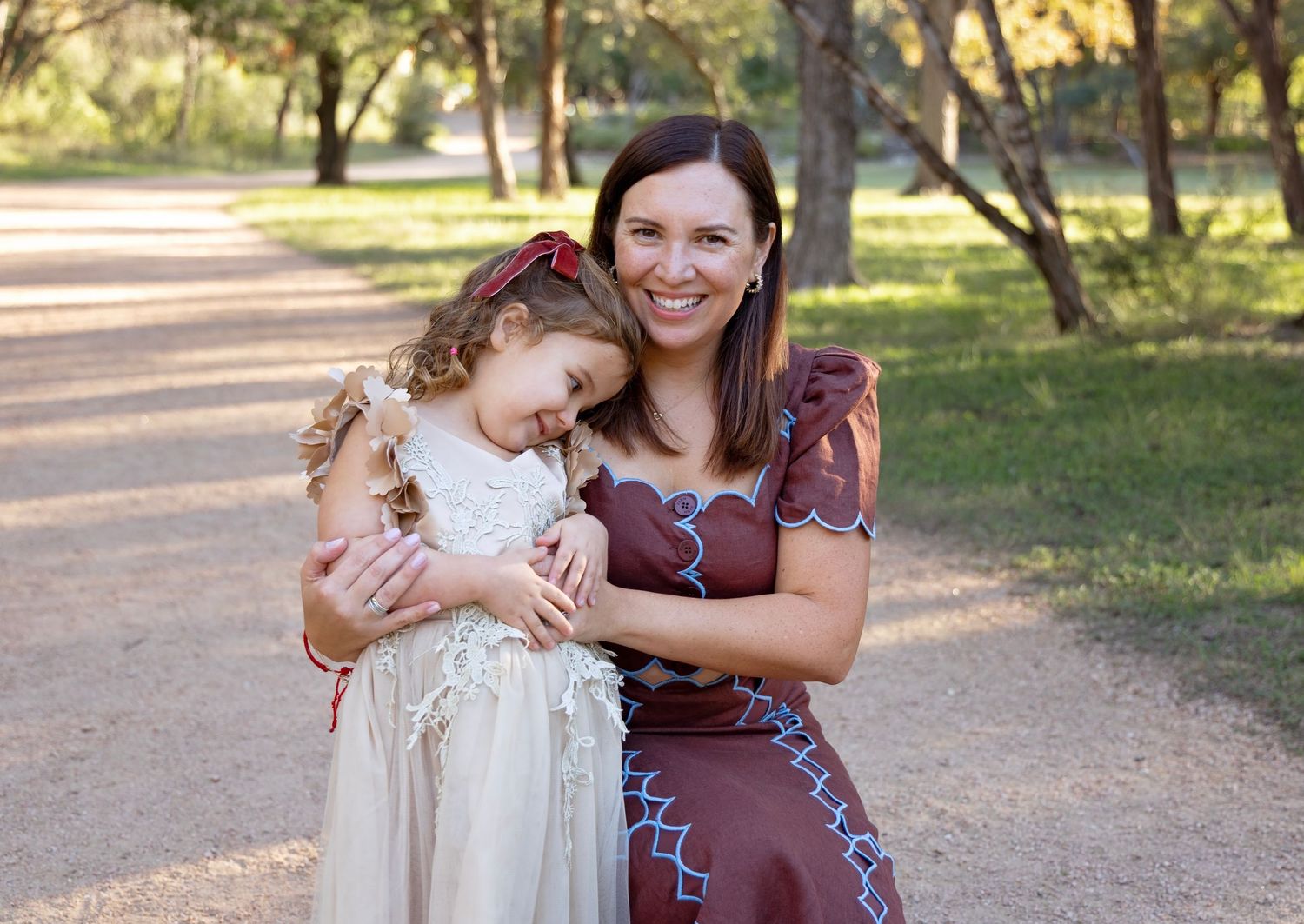 A tender moment shared between two people embracing on a sunlit dirt path surrounded by trees in a park.