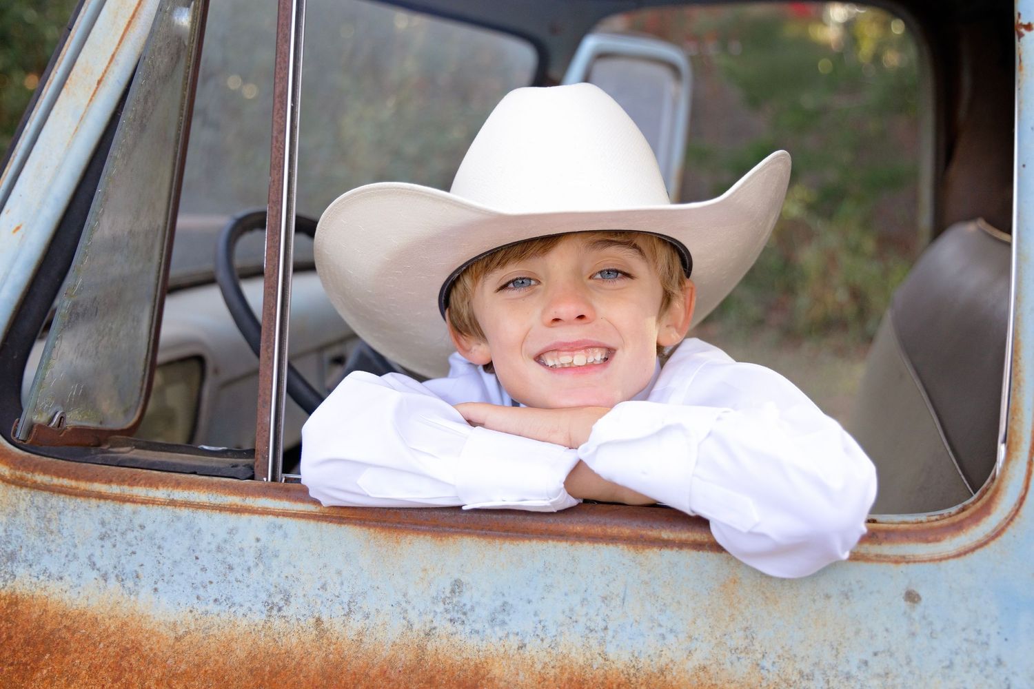 Person wearing a white cowboy hat leans out of a rustic truck window in a rural setting.