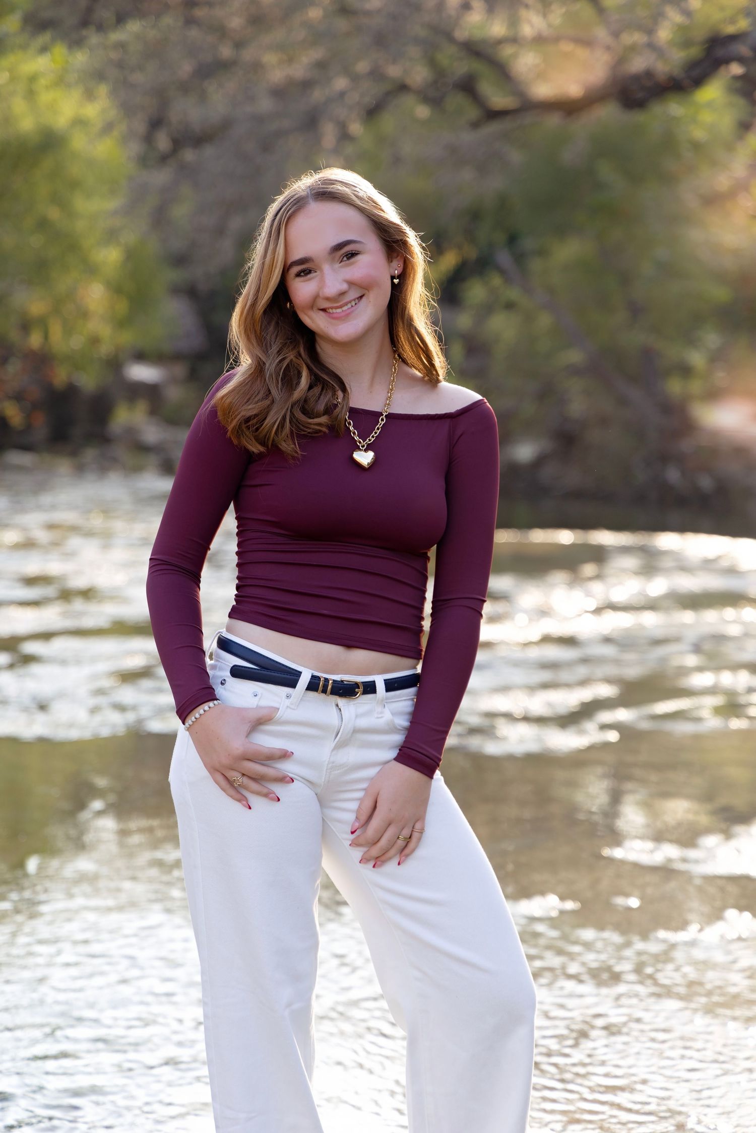 A person in a burgundy long sleeve top and white pants poses outdoors near a river on a sunny day.