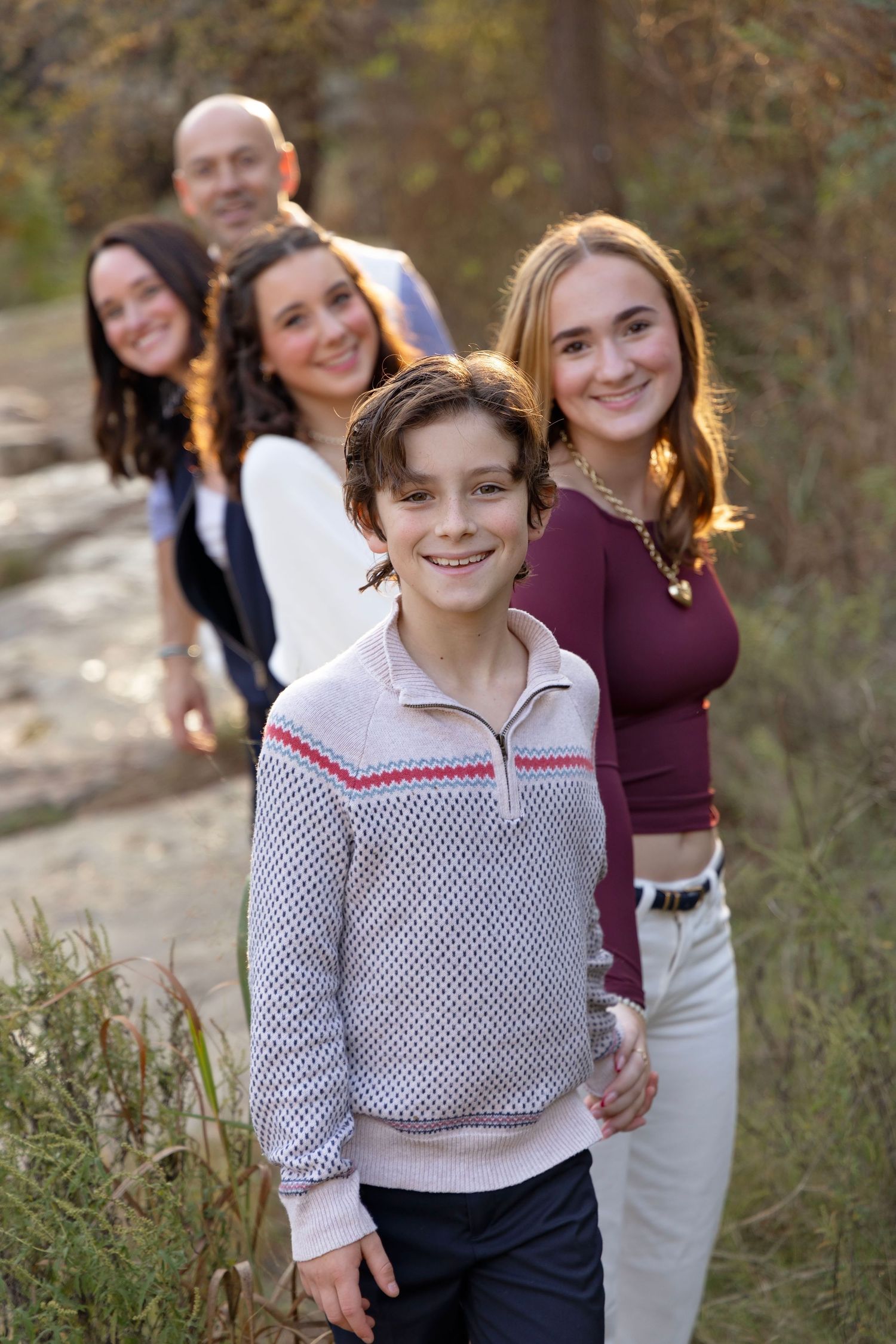 A family poses together outdoors in a line on a nature trail during golden hour lighting.