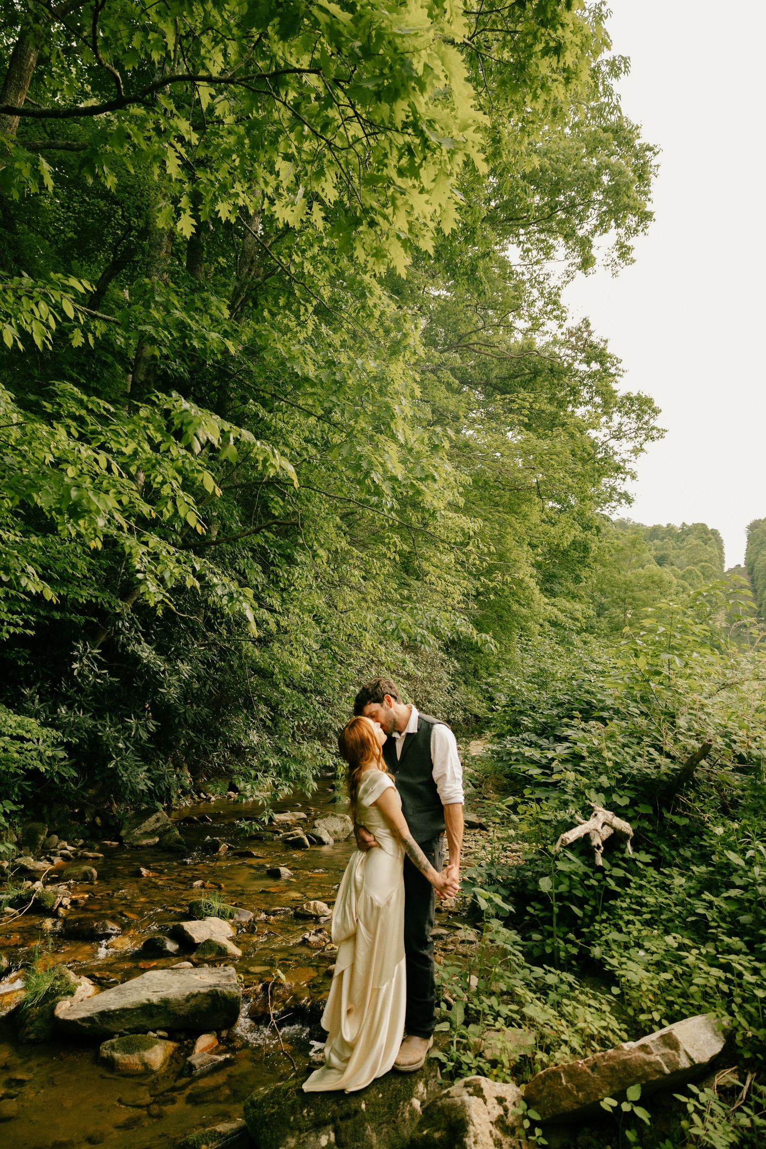 Bride and groom stand on a rock in a creek as they kiss.