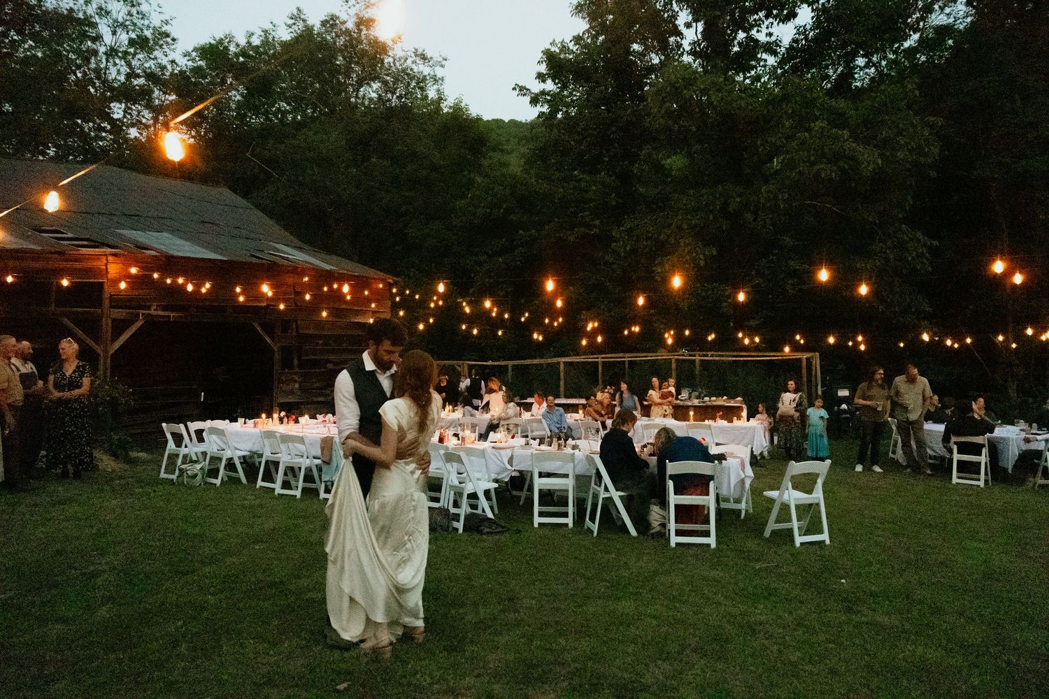 Bride and groom dance outside near the string lights, under the night sky, surrounded by family and loved ones near Asheville, NC.