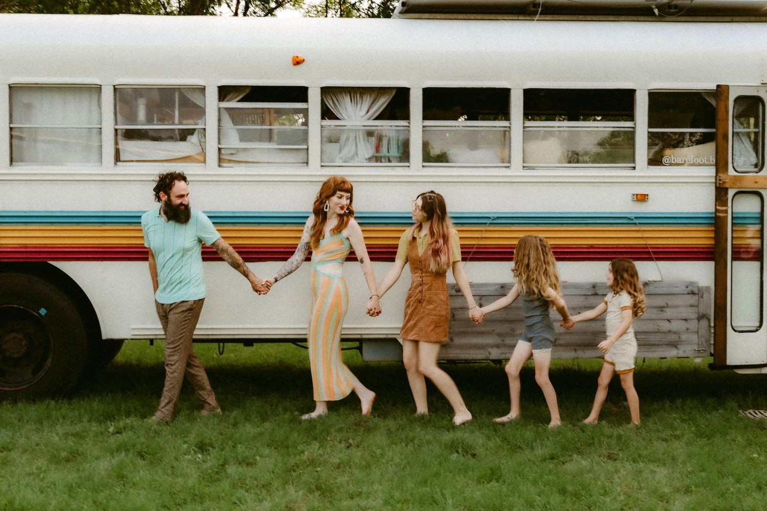 A group holding hands in a line while standing beside a vintage bus with retro rainbow stripes on a grassy lawn.