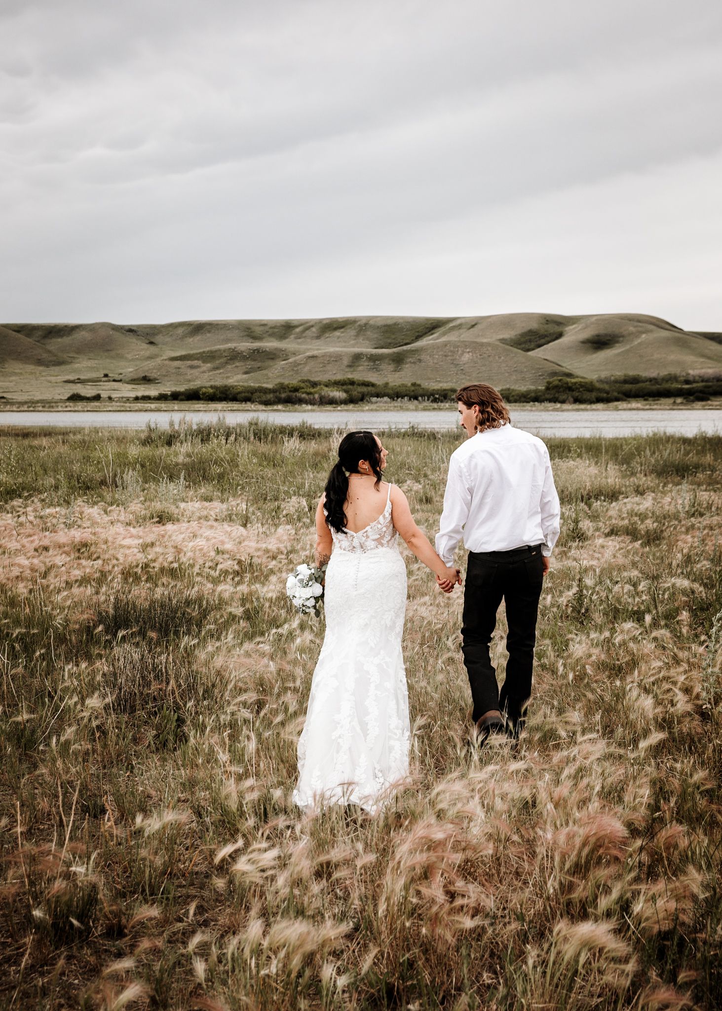 Bride and groom walking hand in hand through a prairie field with rolling hills and a lake in the background. Wedding photography.