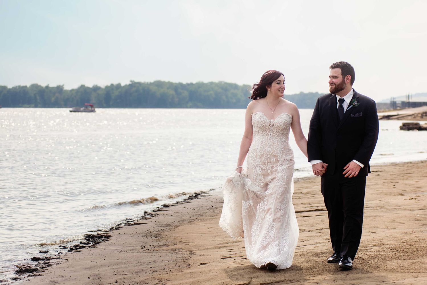 A couple walks along a shoreline in wedding attire with water and boats in the background.
